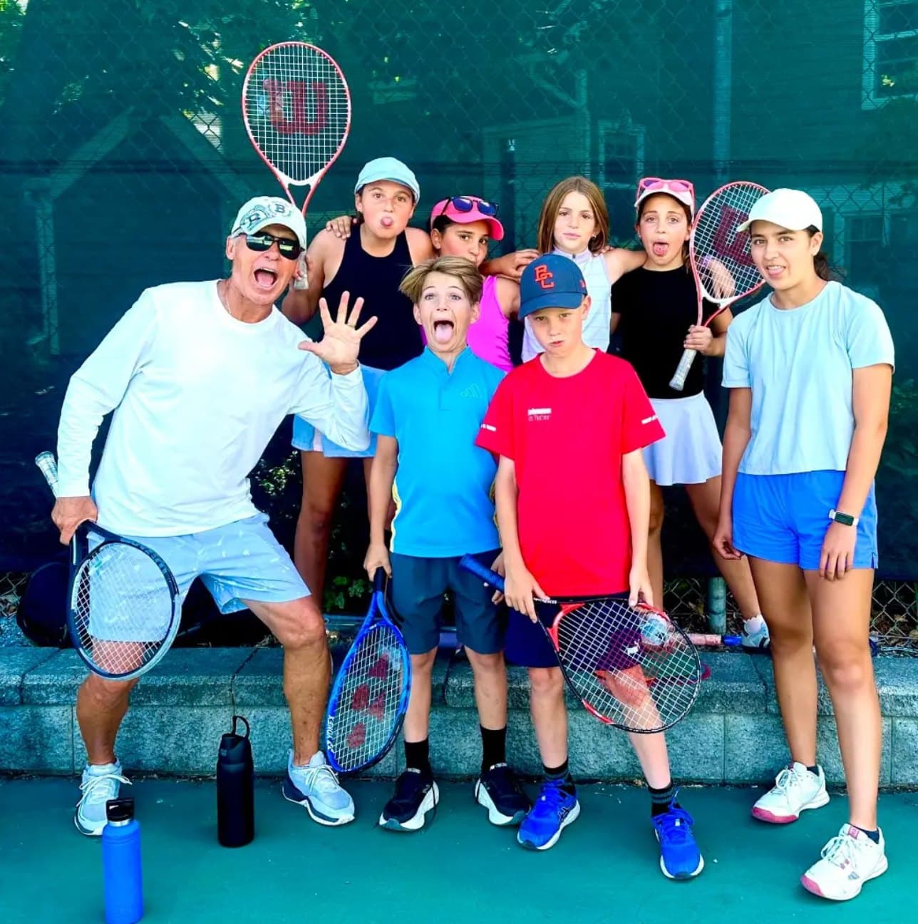 A coach and a group of junior players pulling faces for the camera, racquets in hand, on an outdoor court.